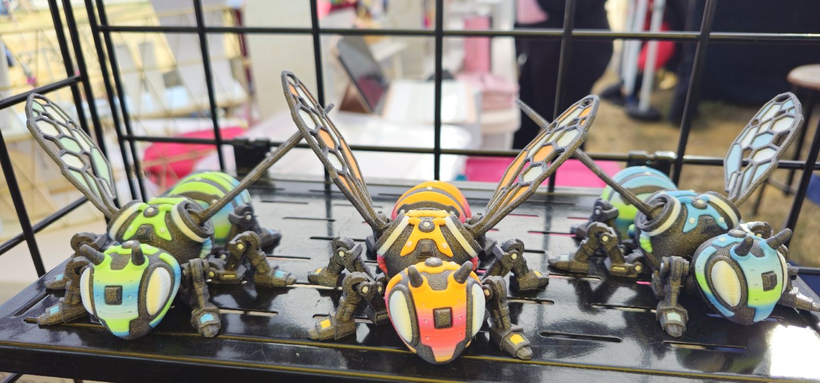 Articulated mechanical bee in orange and pink with honeycomb wings, flanked by green and blue bees